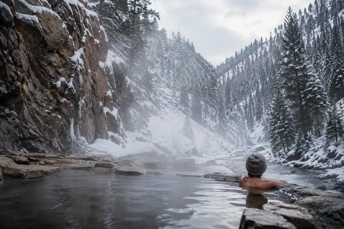 Steam rising from a natural hot spring surrounded by snow with a hiker relaxing during a winter trail hike
