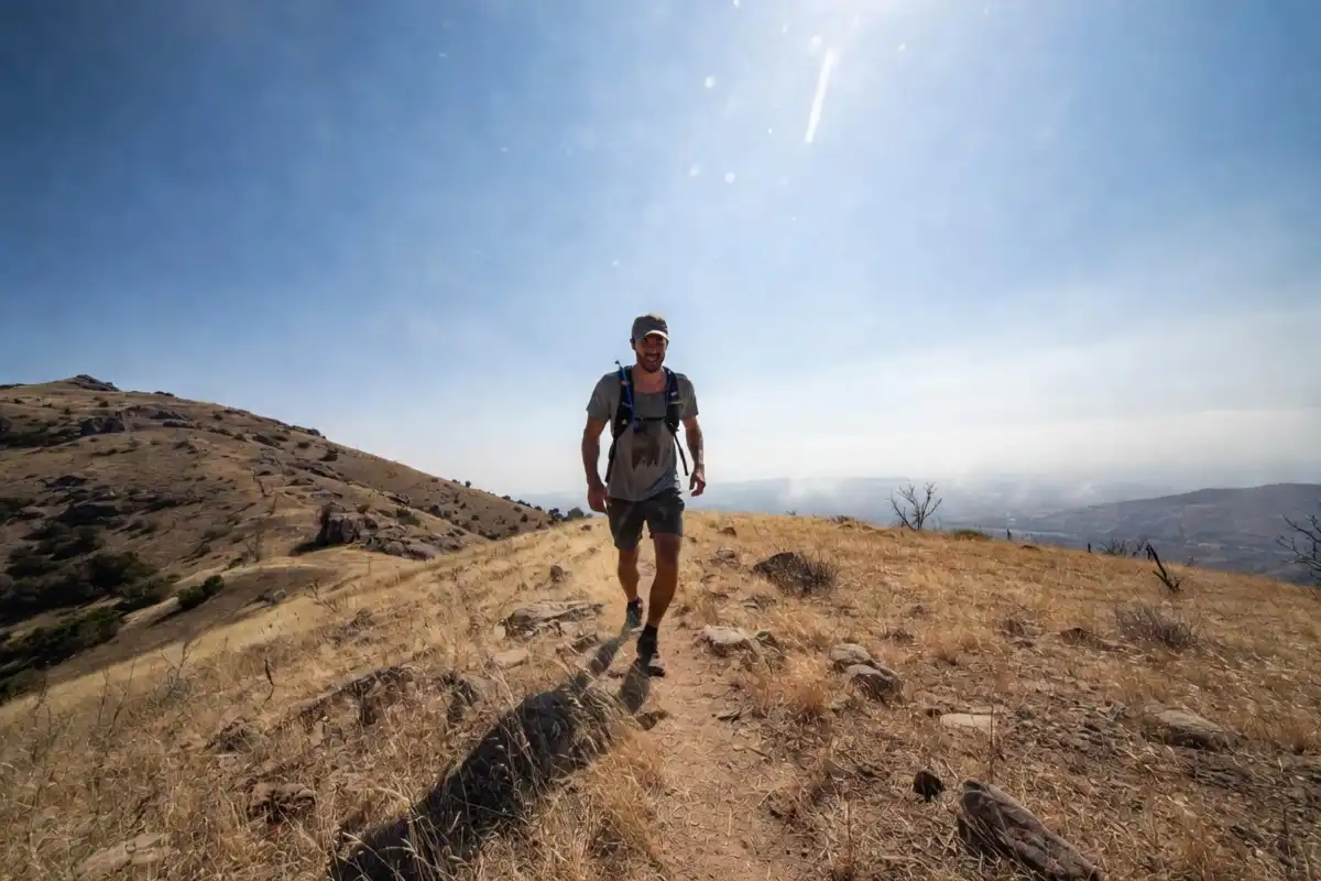 Hiker pausing on an exposed sunny trail wearing a plain cotton shirt with a hydration pack