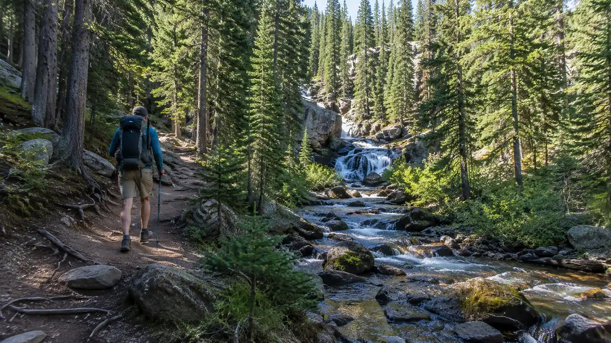 Hiker on Jim Creek Trail near Winter Park, Colorado, walking beside a forest creek toward a cascading waterfall.