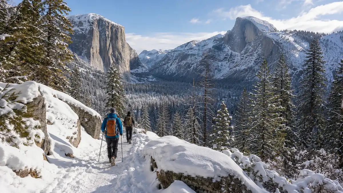 Winter hikers walking on a snowy trail in Yosemite Valley with granite cliffs and pine trees in the background.